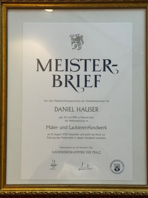 Hand eines Arbeiters in Handschuhen, der eine rote Kelle mit Mörtel oder Putz hält und ihn auf eine graue Wand aufträgt. Nur ein Teil des Arbeiters ist sichtbar, hauptsächlich die Hand und die Kelle. Die Wand im Hintergrund hat eine unebene und unvollendete Textur.